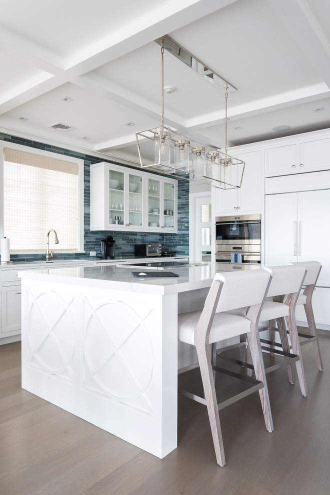 Inspiration for a mid-sized beach style single-wall open plan kitchen in New York with an undermount sink, white cabinets, blue splashback, glass tile splashback, stainless steel appliances, medium hardwood floors, with island, brown floor, white benchtop and shaker cabinets.