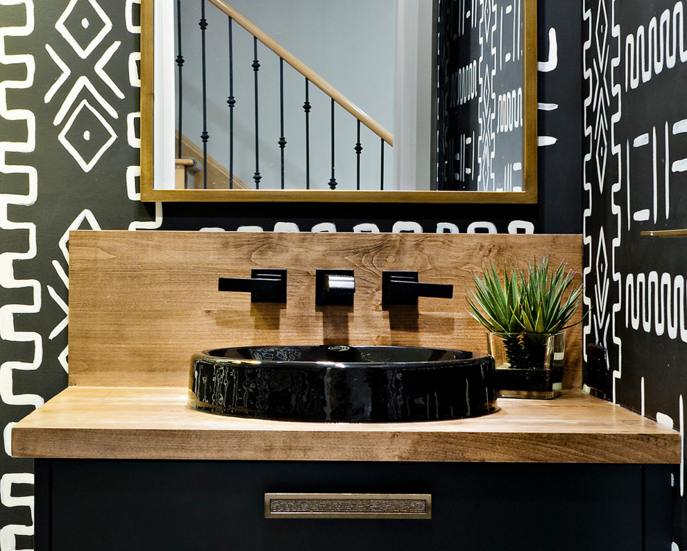 Photo of a mid-sized eclectic powder room in Toronto with flat-panel cabinets, black cabinets, a one-piece toilet, black walls, porcelain floors, a vessel sink, wood benchtops, white floor, brown benchtops, a built-in vanity and wallpaper.