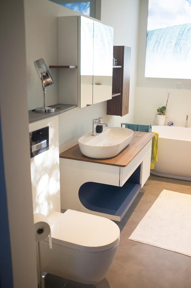 Photo of a mid-sized contemporary master bathroom in Dusseldorf with flat-panel cabinets, dark wood cabinets, a freestanding tub, a two-piece toilet, white tile, white walls, concrete floors, a vessel sink, wood benchtops, grey floor and brown benchtops.