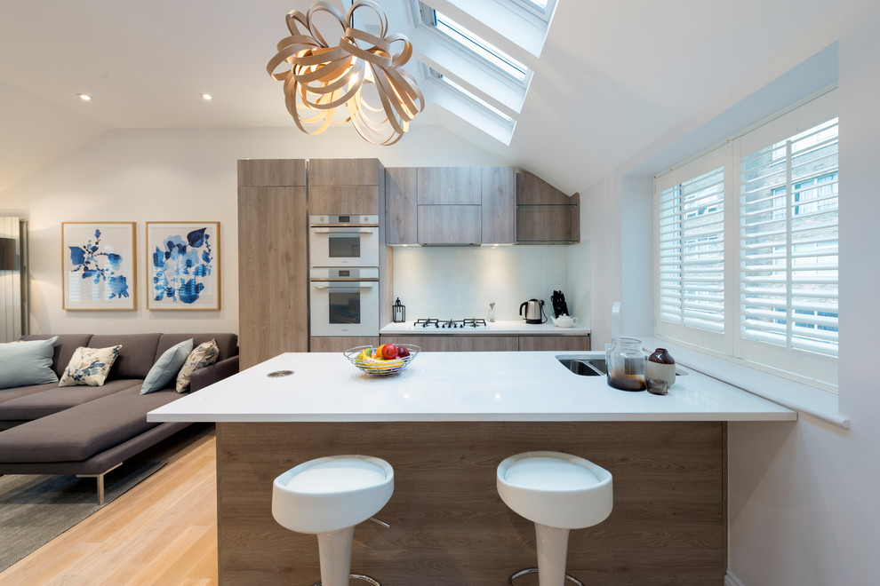 This is an example of a small contemporary galley open plan kitchen in London with flat-panel cabinets, quartzite benchtops, white appliances, light hardwood floors, beige floor, an undermount sink, medium wood cabinets and a peninsula.