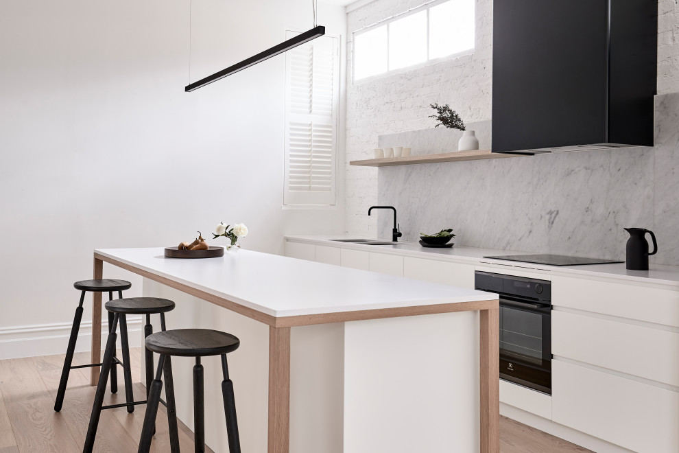 Inspiration for a contemporary galley kitchen in Melbourne with an undermount sink, flat-panel cabinets, white cabinets, grey splashback, stone slab splashback, black appliances, light hardwood floors, with island, beige floor and white benchtop.