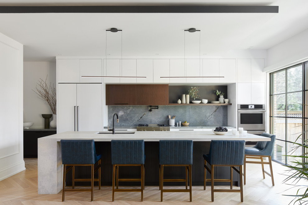 Mid-sized contemporary single-wall open plan kitchen in Toronto with a single-bowl sink, flat-panel cabinets, white cabinets, limestone benchtops, grey splashback, stone slab splashback, stainless steel appliances, light hardwood floors, with island, beige floor, grey benchtop and coffered.