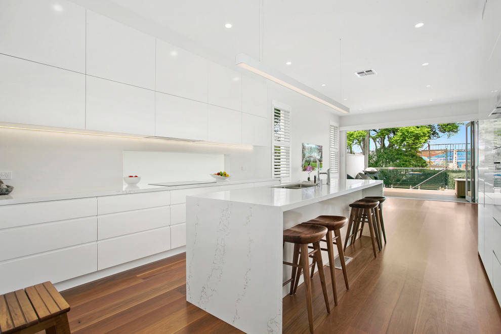 This is an example of a beach style kitchen in Sydney with a double-bowl sink, flat-panel cabinets, white cabinets, white splashback, medium hardwood floors, with island and white benchtop.