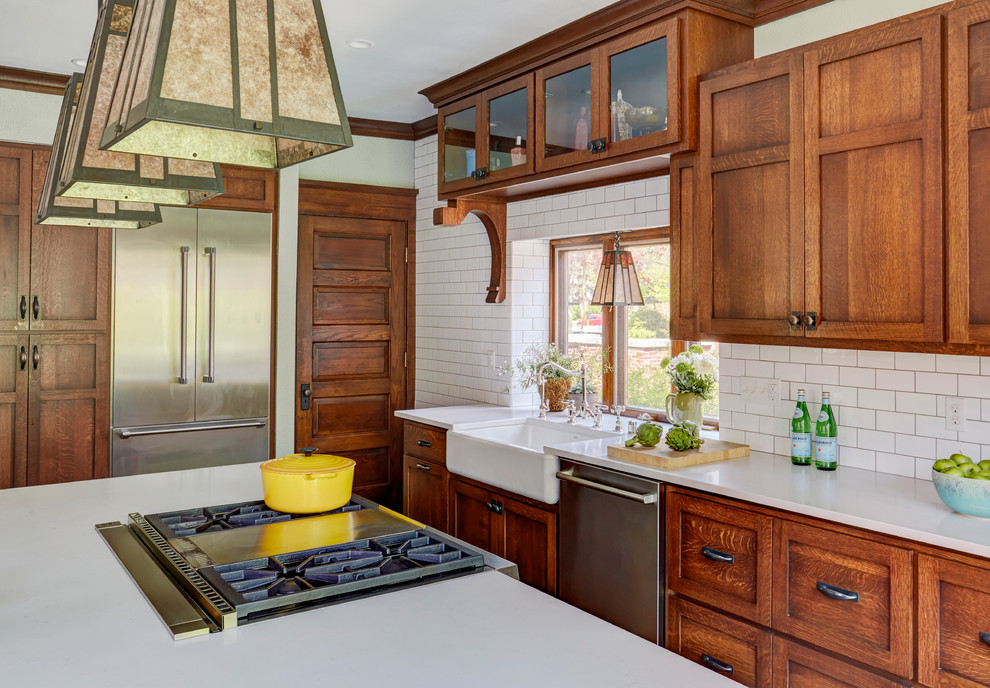 Photo of a mid-sized arts and crafts kitchen in Milwaukee with a farmhouse sink, shaker cabinets, medium wood cabinets, quartz benchtops, white splashback, subway tile splashback, stainless steel appliances, medium hardwood floors, with island, brown floor and white benchtop.