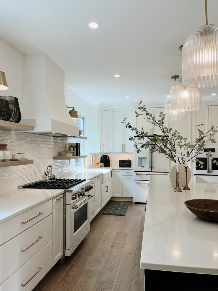 Eat-in kitchen - large transitional l-shaped light wood floor and beige floor eat-in kitchen idea with a farmhouse sink, shaker cabinets, white cabinets, quartz countertops, white backsplash, ceramic backsplash, white appliances, an island and white countertops