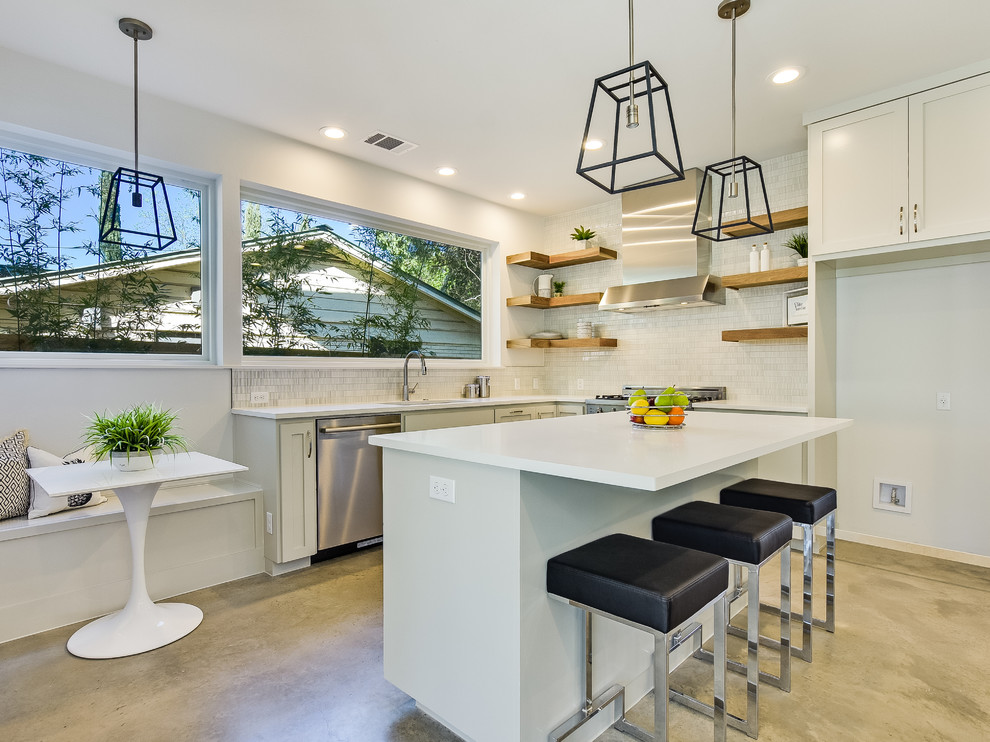 Photo of a mid-sized midcentury single-wall open plan kitchen in Austin with an undermount sink, shaker cabinets, white cabinets, quartz benchtops, white splashback, stone tile splashback, stainless steel appliances, concrete floors, with island and brown floor.