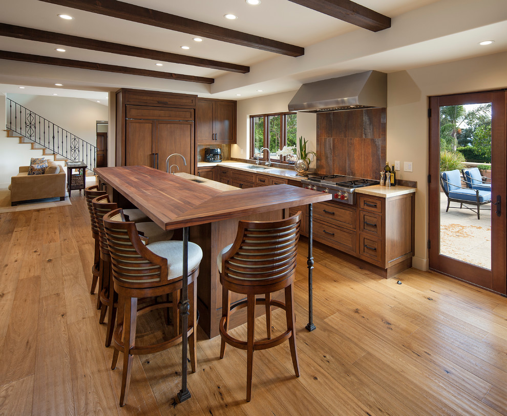 Photo of a mid-sized mediterranean galley eat-in kitchen in Santa Barbara with a double-bowl sink, recessed-panel cabinets, brown cabinets, wood benchtops, brown splashback, ceramic splashback, panelled appliances, light hardwood floors and with island.
