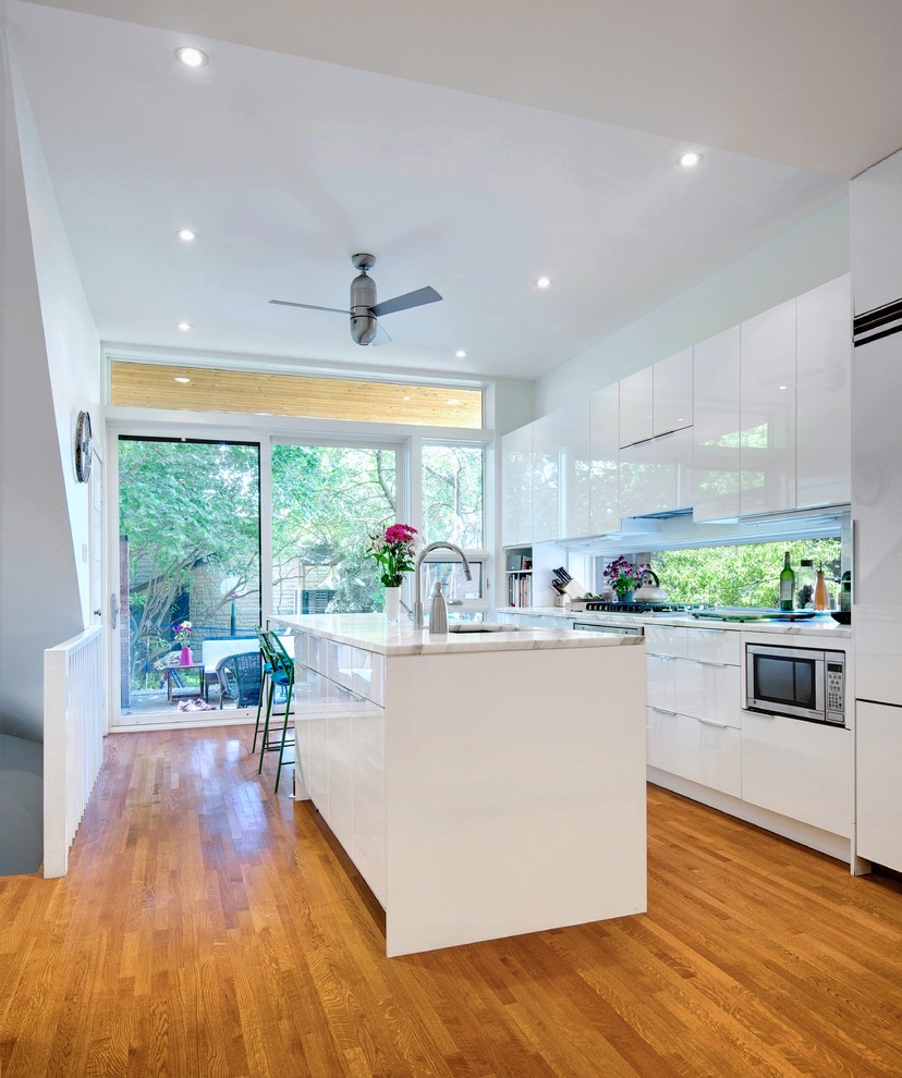 Inspiration for a large contemporary galley separate kitchen in Toronto with a double-bowl sink, flat-panel cabinets, white cabinets, marble benchtops, white splashback, medium hardwood floors, with island, window splashback and panelled appliances.