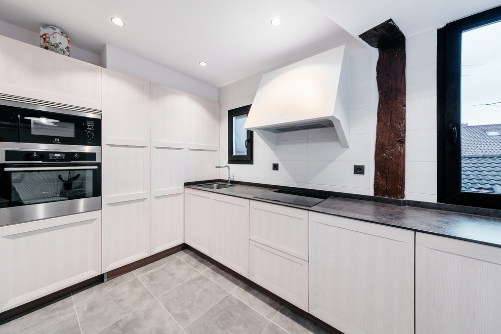 Photo of a contemporary l-shaped kitchen in Bilbao with an undermount sink, beaded inset cabinets, light wood cabinets, white splashback, stainless steel appliances, no island, grey floor and black benchtop.