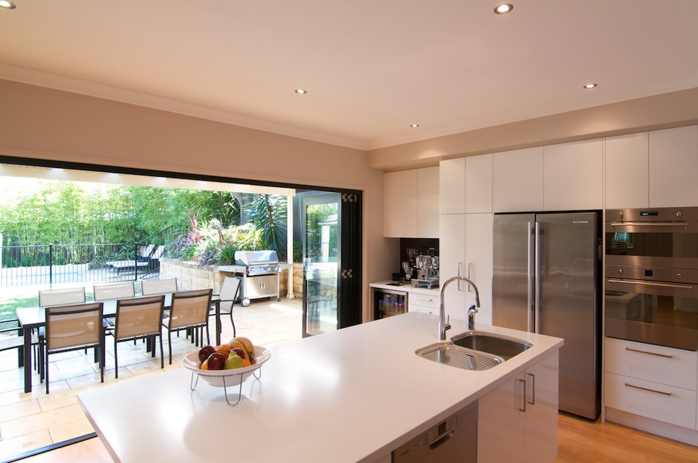 This is an example of a mid-sized modern l-shaped eat-in kitchen in Sydney with a triple-bowl sink, white cabinets, glass sheet splashback, stainless steel appliances, with island, flat-panel cabinets, solid surface benchtops, black splashback and light hardwood floors.
