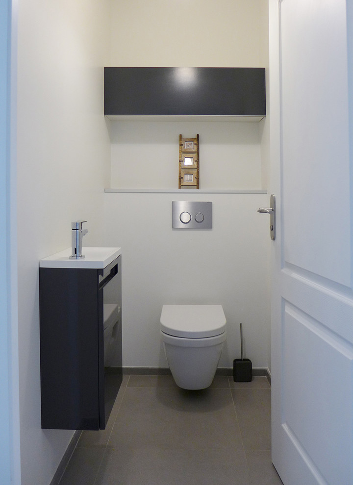 Photo of a mid-sized contemporary powder room in Montpellier with beaded inset cabinets, grey cabinets, a wall-mount toilet, white tile, white walls, ceramic floors, a wall-mount sink, glass benchtops, grey floor and white benchtops.
