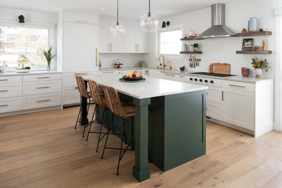 This is an example of a transitional l-shaped open plan kitchen in Denver with a farmhouse sink, shaker cabinets, green cabinets, quartzite benchtops, white splashback, ceramic splashback, stainless steel appliances, light hardwood floors, with island, white benchtop and brown floor.