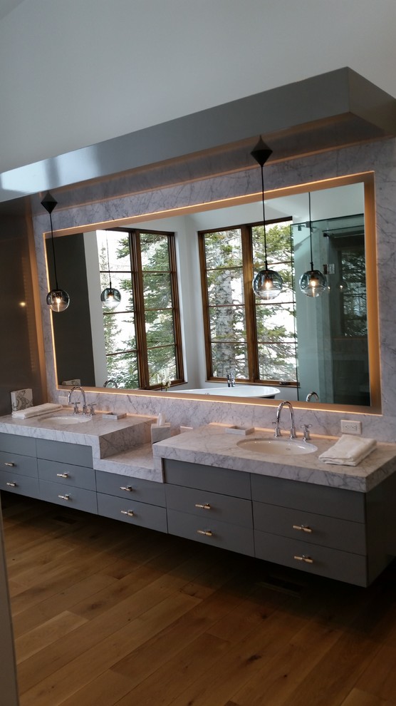 Photo of a large modern master bathroom in Salt Lake City with grey walls, medium hardwood floors, an undermount sink, brown floor, flat-panel cabinets, grey cabinets, a freestanding tub, a corner shower, marble benchtops, a hinged shower door and white benchtops.