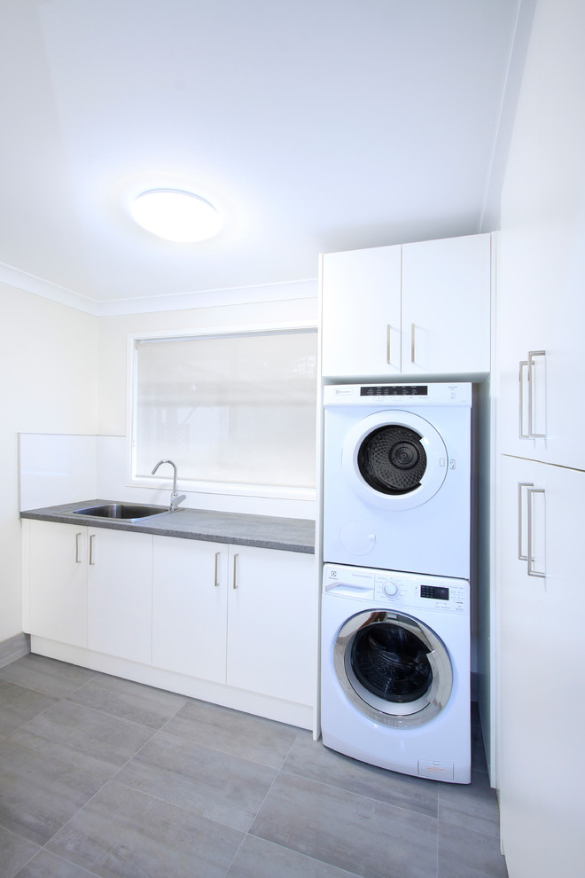 Mid-sized modern l-shaped dedicated laundry room in Wollongong with a drop-in sink, flat-panel cabinets, white cabinets, laminate benchtops, white walls, ceramic floors, a stacked washer and dryer, grey floor and grey benchtop.