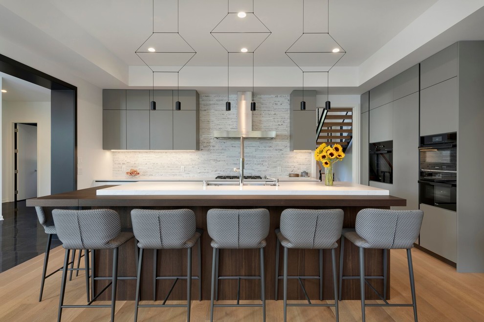 Design ideas for a contemporary l-shaped kitchen in Minneapolis with an undermount sink, flat-panel cabinets, grey cabinets, grey splashback, black appliances, medium hardwood floors, with island, brown floor and white benchtop.