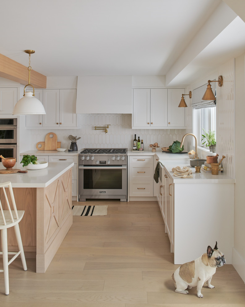 Large farmhouse l-shaped kitchen pantry in Toronto with a belfast sink, shaker cabinets, white cabinets, engineered stone countertops, white splashback, ceramic splashback, integrated appliances, light hardwood flooring, an island, white worktops and exposed beams.