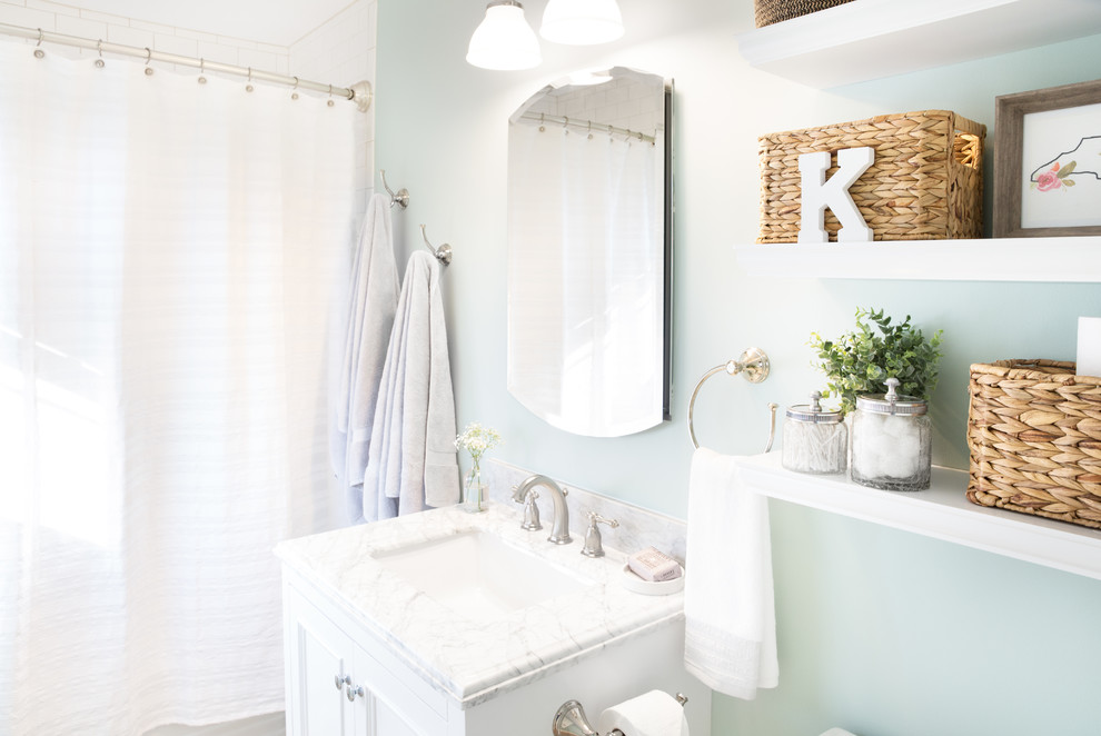 Design ideas for a mid-sized beach style bathroom in Charlotte with raised-panel cabinets, white cabinets, an alcove tub, a shower/bathtub combo, a one-piece toilet, white tile, ceramic tile, blue walls, porcelain floors, an undermount sink, marble benchtops, brown floor and a shower curtain.