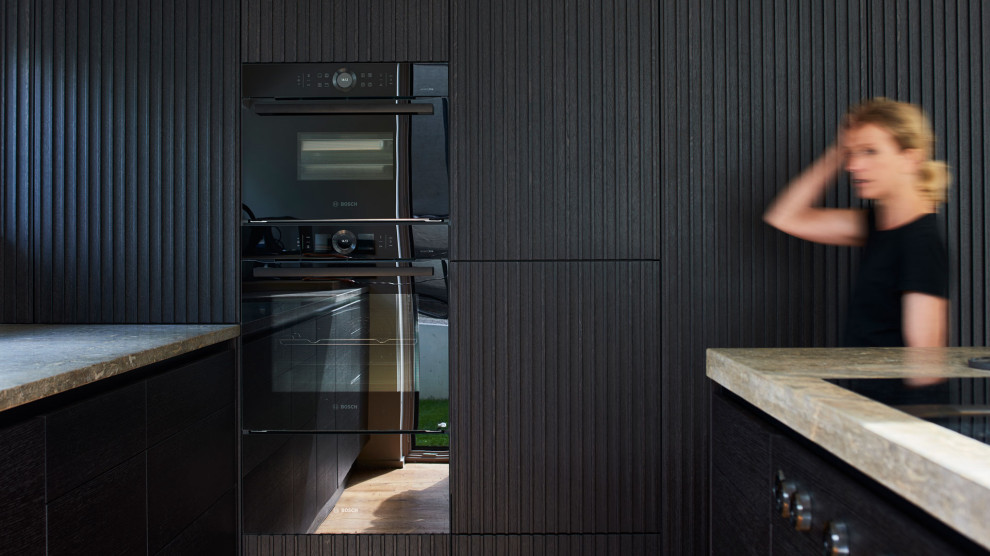 This is an example of a contemporary kitchen in Munich with louvered cabinets, black cabinets, granite benchtops, black appliances, painted wood floors, with island, brown floor and beige benchtop.