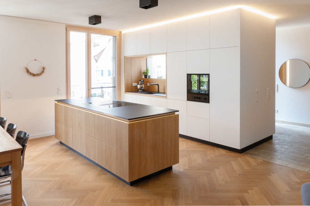 Küche With flächenbündigen Schrankfronten, weißen Schränken, schwarzen Elektrogeräten, braunem Holzboden, Kücheninsel, braunem Boden and schwarzer Arbeitsplatte in Stuttgart
