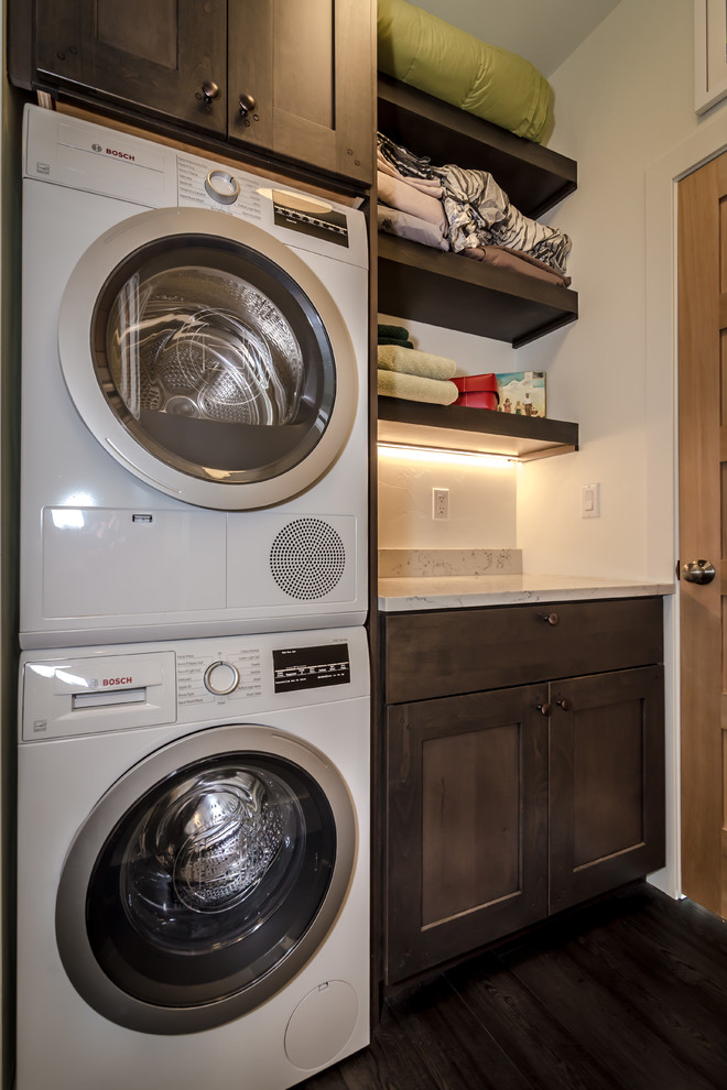 Small modern single-wall dedicated laundry room in Denver with shaker cabinets, dark wood cabinets, quartz benchtops, white walls, laminate floors, a stacked washer and dryer, brown floor and white benchtop.
