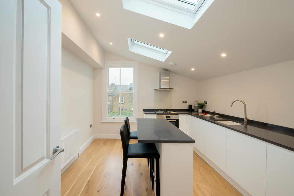 This is an example of a contemporary l-shaped kitchen in London with an undermount sink, flat-panel cabinets, stainless steel appliances, light hardwood floors, with island, beige floor and black benchtop.