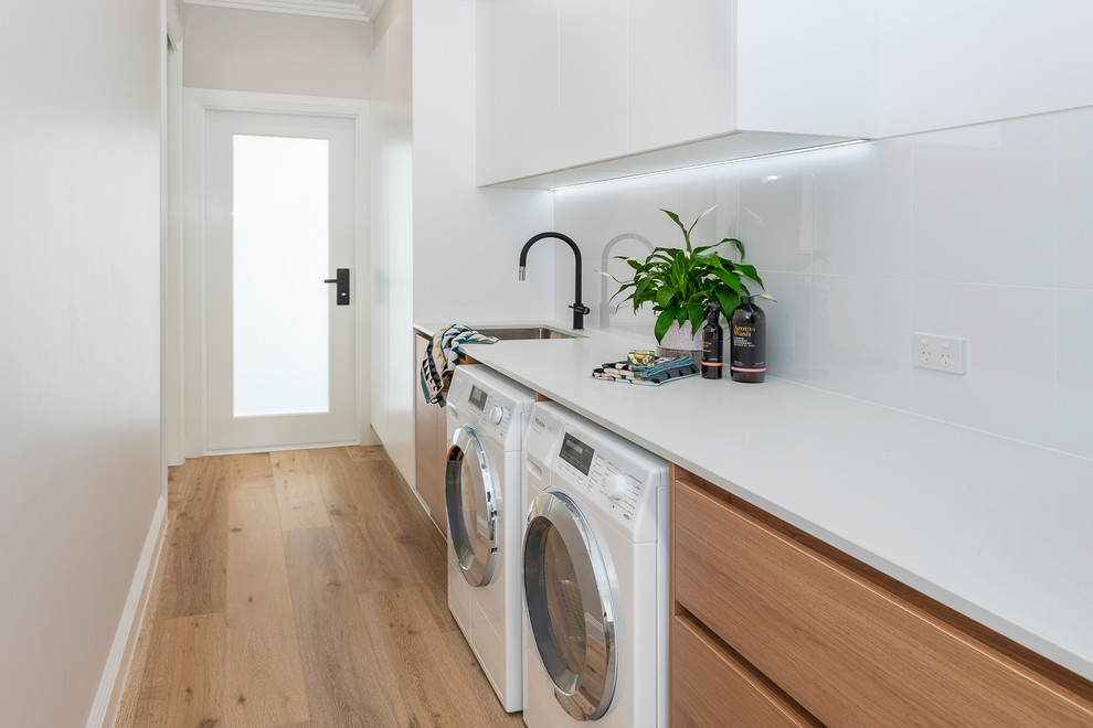 Large contemporary galley dedicated laundry room in Brisbane with an undermount sink, light wood cabinets, quartz benchtops, white walls, light hardwood floors, a side-by-side washer and dryer and white benchtop.