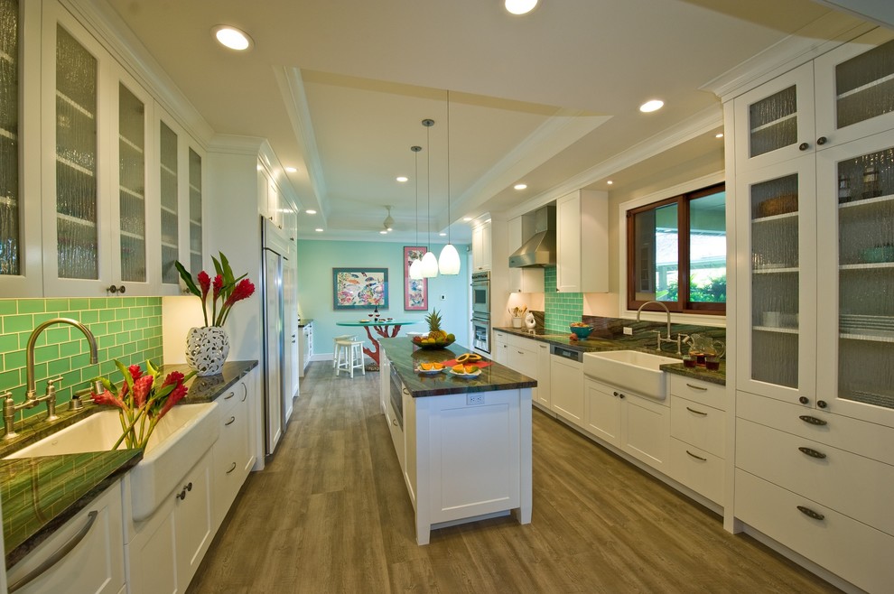 This is an example of a tropical kitchen in Hawaii with a farmhouse sink, shaker cabinets, white cabinets, granite benchtops, green splashback, glass tile splashback, panelled appliances, vinyl floors and with island.