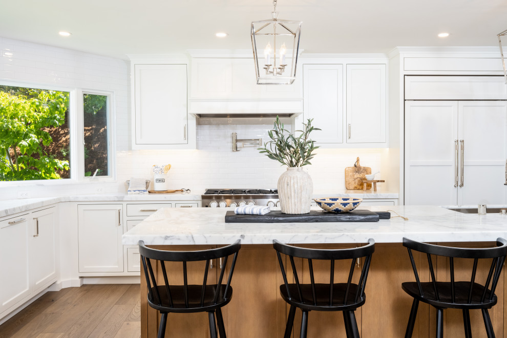 This is an example of a large country l-shaped open plan kitchen in San Francisco with an undermount sink, shaker cabinets, white cabinets, marble benchtops, white splashback, ceramic splashback, panelled appliances, medium hardwood floors, with island, brown floor and white benchtop.