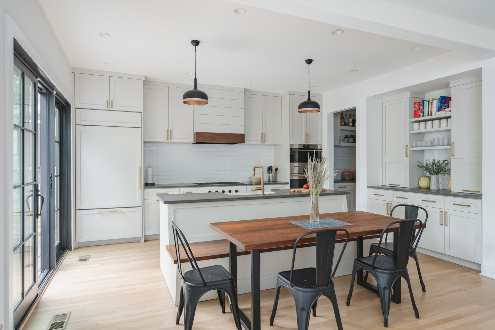 This is an example of a classic l-shaped kitchen/diner in DC Metro with a belfast sink, shaker cabinets, white cabinets, white splashback, metro tiled splashback, integrated appliances, light hardwood flooring, an island, beige floors and grey worktops.