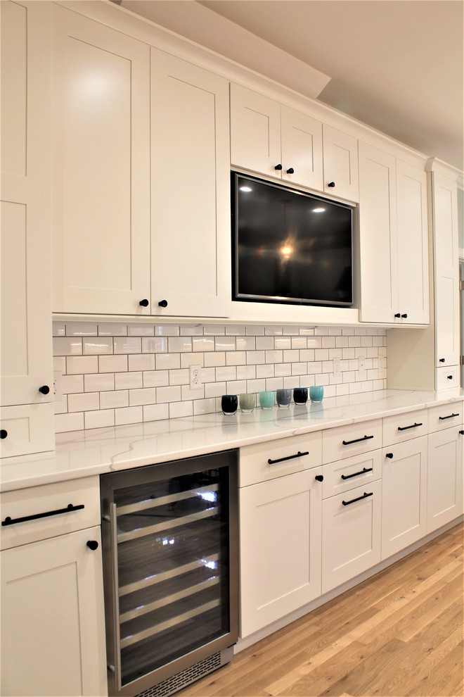 Large transitional galley light wood floor eat-in kitchen photo with a farmhouse sink, shaker cabinets, white cabinets, quartz countertops, white backsplash, ceramic backsplash, stainless steel appliances, an island and white countertops