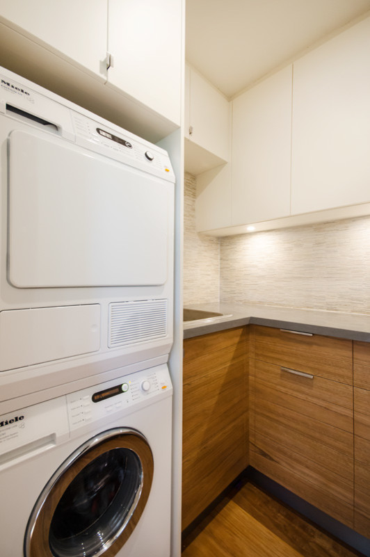 Inspiration for a small contemporary u-shaped utility room in Melbourne with a drop-in sink, flat-panel cabinets, medium wood cabinets, quartz benchtops, beige walls, plywood floors and a stacked washer and dryer.