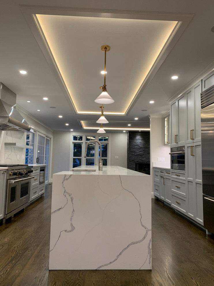 Large transitional galley dark wood floor and brown floor enclosed kitchen photo in Boston with a farmhouse sink, raised-panel cabinets, quartzite countertops, white backsplash, stone slab backsplash, stainless steel appliances, an island, white countertops and gray cabinets