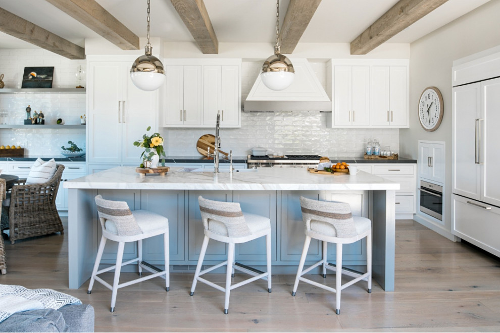 Mid-sized beach style open plan kitchen in Orange County with a farmhouse sink, shaker cabinets, white cabinets, marble benchtops, white splashback, ceramic splashback, panelled appliances, light hardwood floors, with island, beige floor, white benchtop and exposed beam.