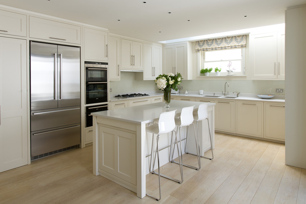 Mid-sized traditional l-shaped kitchen in London with a double-bowl sink, shaker cabinets, white cabinets, white splashback, glass sheet splashback, stainless steel appliances, light hardwood floors and with island.