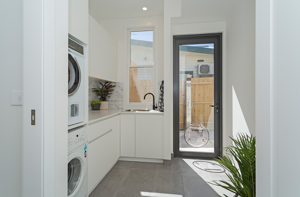 Mid-sized modern l-shaped dedicated laundry room in Other with white cabinets, white walls, concrete floors, grey floor, white benchtop, a stacked washer and dryer, a single-bowl sink, flat-panel cabinets, multi-coloured splashback and ceramic splashback.
