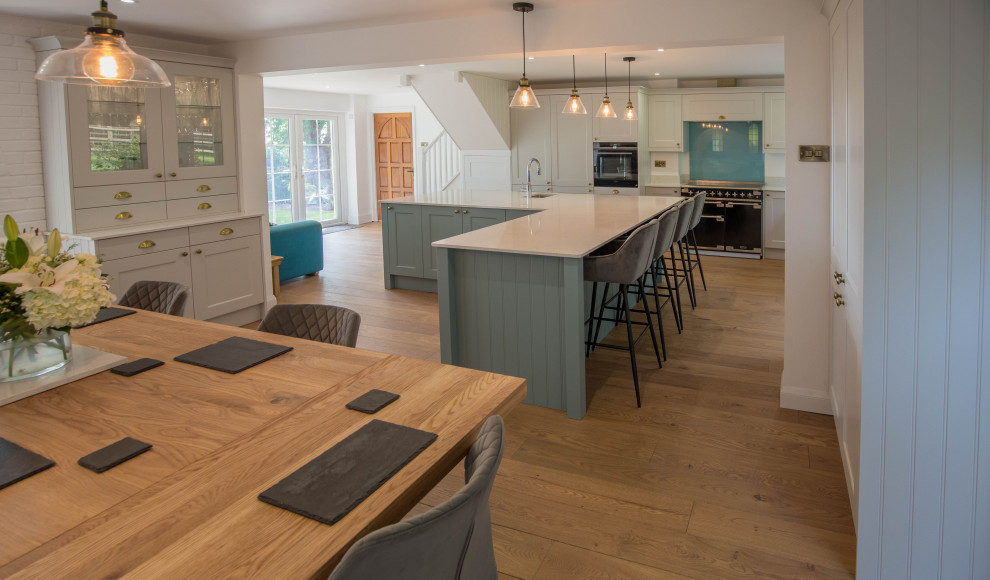 Design ideas for a large contemporary l-shaped open plan kitchen in Devon with a farmhouse sink, shaker cabinets, turquoise cabinets, quartzite benchtops, blue splashback, glass sheet splashback, black appliances, light hardwood floors, with island, brown floor and white benchtop.