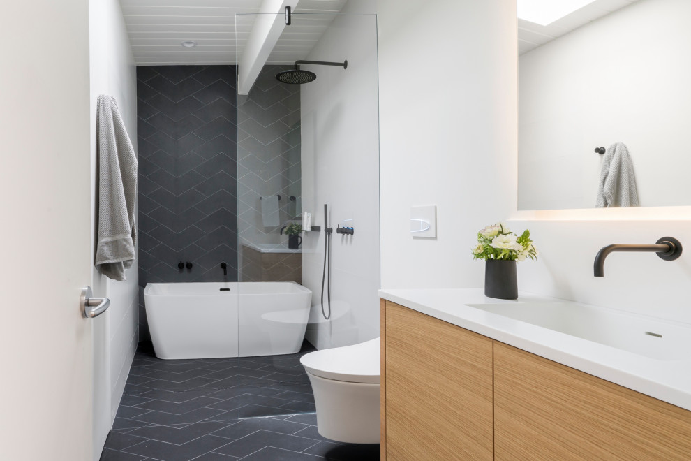 Inspiration for a contemporary wet room bathroom in San Francisco with flat-panel cabinets, light wood cabinets, a freestanding bath, a wall mounted toilet, grey tiles, ceramic tiles, white walls, ceramic flooring, engineered stone worktops, grey floors, white worktops, a single sink and exposed beams.