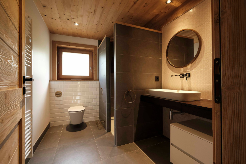Foto på ett litet rustikt brun badrum med dusch, med bruna skåp, en kantlös dusch, en vägghängd toalettstol, rosa kakel, cementkakel, vita väggar, klinkergolv i keramik, ett avlångt handfat, träbänkskiva, grått golv och med dusch som är öppen
