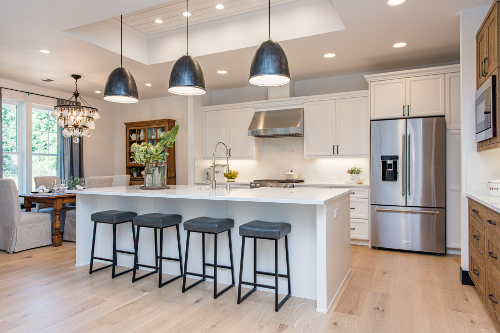 Transitional l-shaped open plan kitchen in Seattle with a farmhouse sink, recessed-panel cabinets, white cabinets, quartz benchtops, white splashback, stainless steel appliances, light hardwood floors, with island, brown floor and white benchtop.