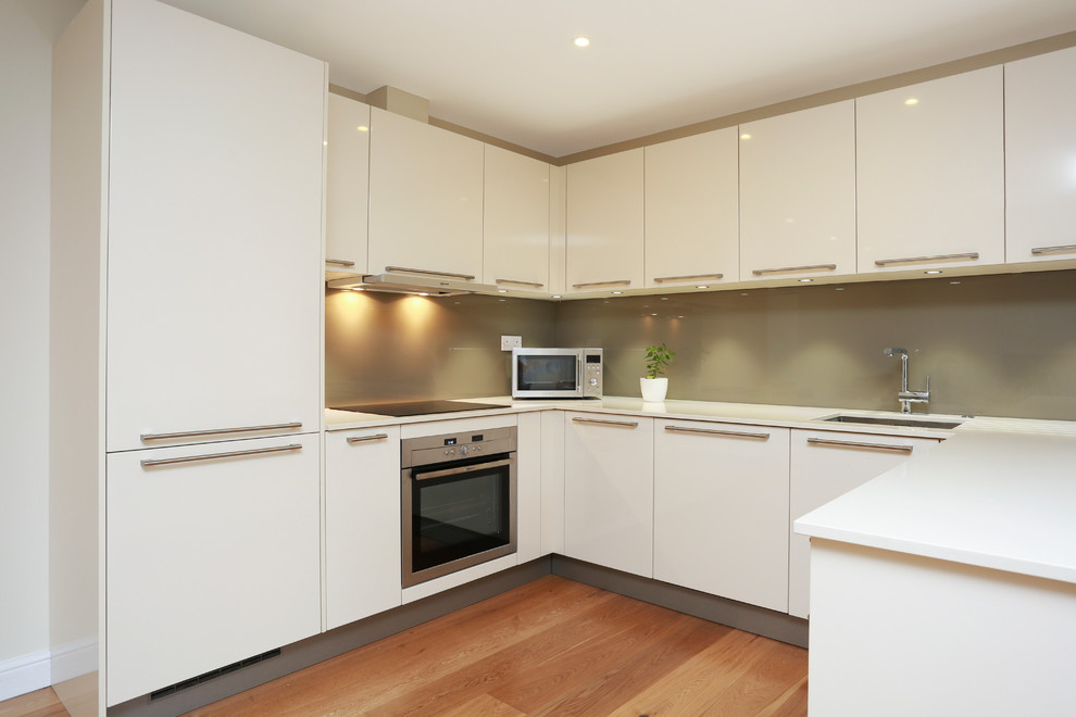Photo of a mid-sized modern u-shaped open plan kitchen in London with green splashback, glass sheet splashback, stainless steel appliances, a peninsula and flat-panel cabinets.