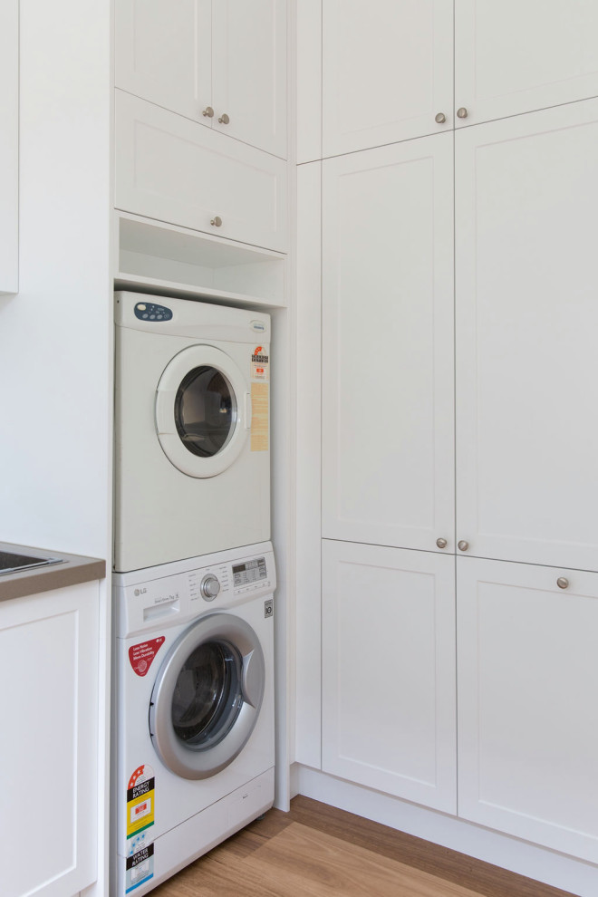 Mid-sized traditional l-shaped utility room in Melbourne with a double-bowl sink, shaker cabinets, white cabinets, quartz benchtops, white walls, medium hardwood floors and a stacked washer and dryer.