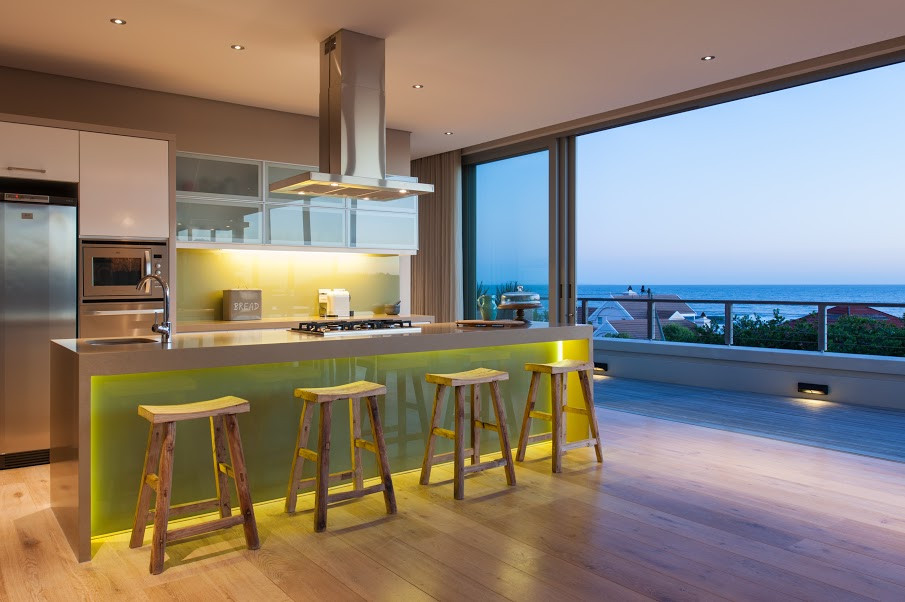 Photo of a mid-sized beach style open plan kitchen in Other with a double-bowl sink, glass-front cabinets, beige cabinets, quartz benchtops, green splashback, glass sheet splashback, stainless steel appliances, light hardwood floors, with island, beige floor and beige benchtop.