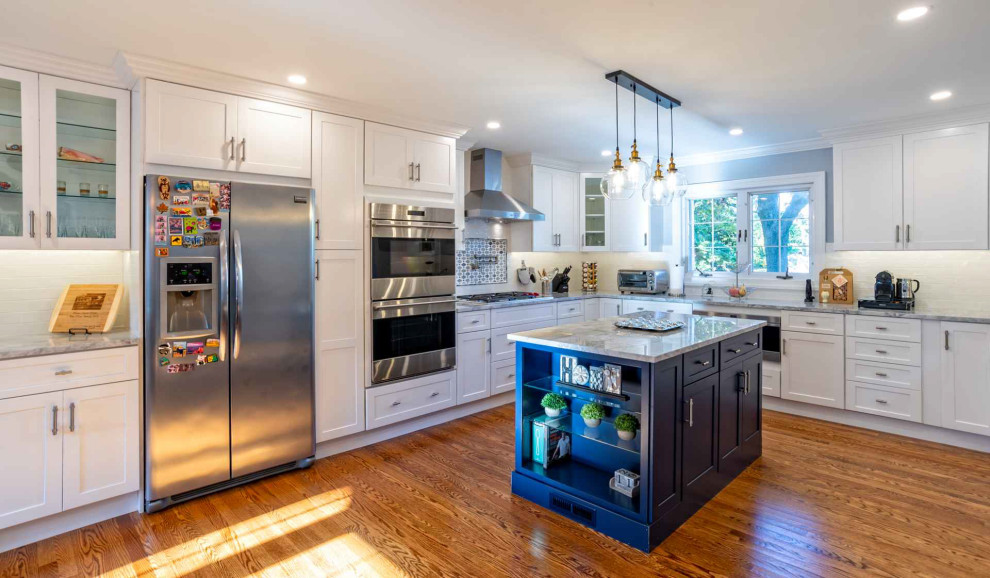 This is an example of a mid-sized transitional u-shaped eat-in kitchen in Philadelphia with a farmhouse sink, shaker cabinets, blue cabinets, quartz benchtops, white splashback, porcelain splashback, stainless steel appliances, medium hardwood floors, with island, brown floor and grey benchtop.