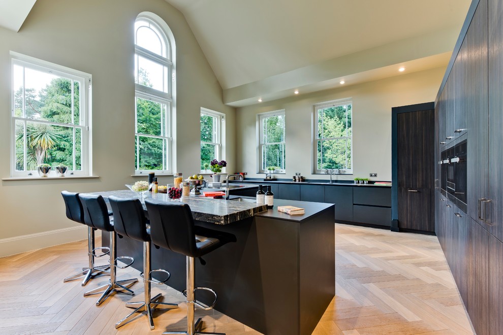 This is an example of a large contemporary u-shaped kitchen in Surrey with an undermount sink, flat-panel cabinets, black cabinets, light hardwood floors, with island, beige floor, black benchtop, beige splashback and panelled appliances.
