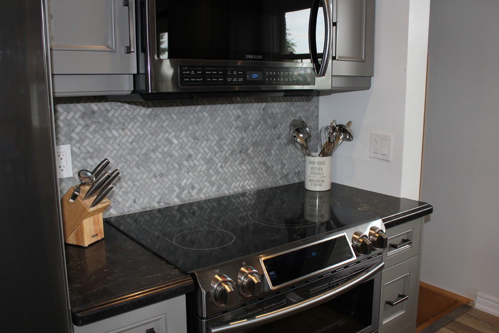 This is an example of a mid-sized transitional l-shaped open plan kitchen in Toronto with beaded inset cabinets, grey cabinets, mosaic tile splashback, black appliances, vinyl floors, with island, an undermount sink, grey splashback, beige floor and black benchtop.