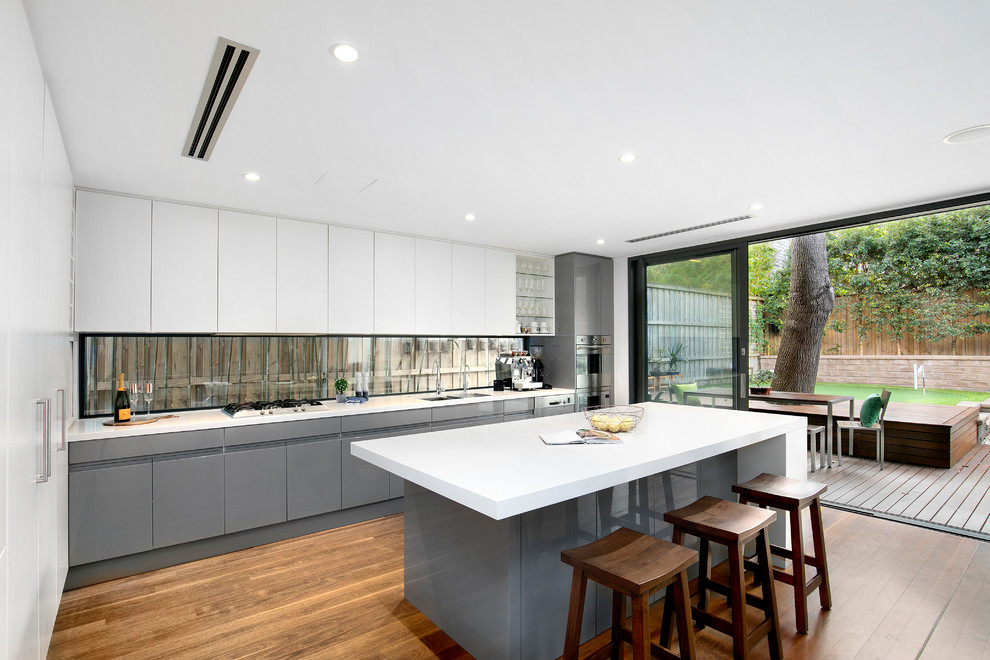 Large modern single-wall kitchen in Sydney with an undermount sink, flat-panel cabinets, grey cabinets, solid surface benchtops, window splashback, stainless steel appliances, medium hardwood floors, with island, brown floor and white benchtop.