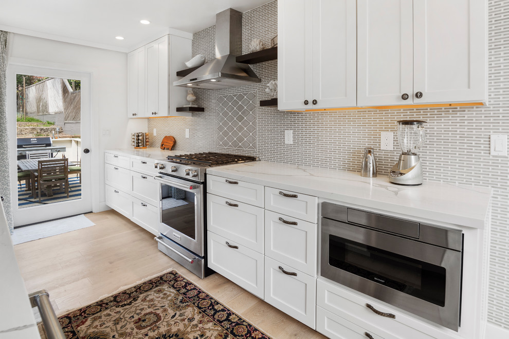 Inspiration for a mid-sized beach style open plan kitchen in San Francisco with an undermount sink, shaker cabinets, white cabinets, quartz benchtops, grey splashback, ceramic splashback, stainless steel appliances, light hardwood floors, with island and white benchtop.
