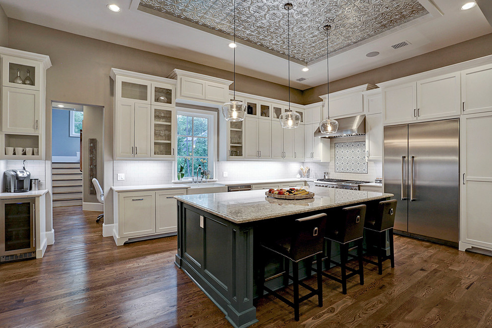 Photo of a large country l-shaped eat-in kitchen in Houston with a farmhouse sink, white cabinets, granite benchtops, white splashback, ceramic splashback, stainless steel appliances, medium hardwood floors, with island, brown floor and shaker cabinets.