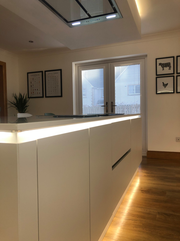 Photo of a large contemporary l-shaped eat-in kitchen in Glasgow with an integrated sink, flat-panel cabinets, white cabinets, quartzite benchtops, green splashback, glass sheet splashback, black appliances, vinyl floors, with island, brown floor, white benchtop and recessed.