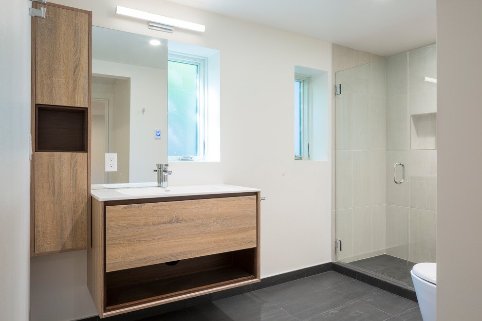 This is an example of a mid-sized contemporary 3/4 bathroom in Seattle with flat-panel cabinets, light wood cabinets, an alcove shower, a one-piece toilet, beige tile, porcelain tile, white walls, porcelain floors, a wall-mount sink, black floor, a hinged shower door and engineered quartz benchtops.